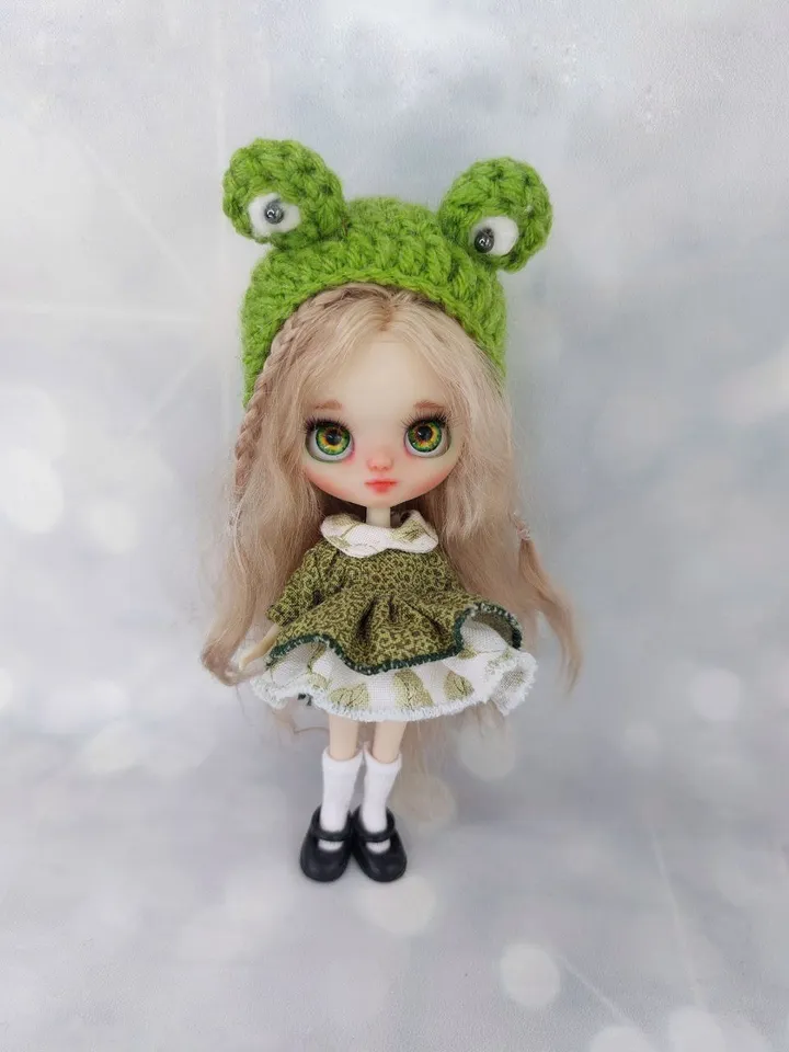 A miniature Blythe doll with wavy blonde hair. The frog cap.