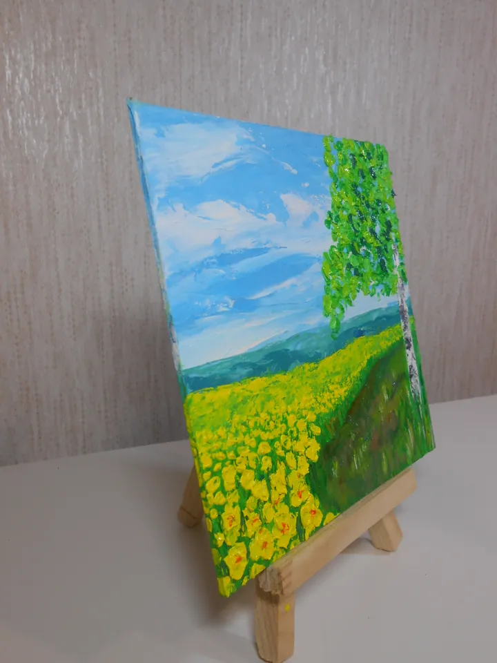A bright landscape. Mountains in the distance, blue sky with light clouds, a field of yellow flowers, a birch tree. The original painting.